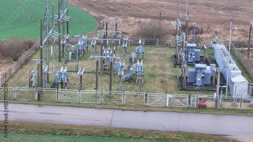 A fenced rural electrical substation surrounded by power lines, transformers, open agricultural fields and distant forest under an overcast sky, expressing industrial presence in natural countryside.