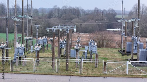 A fenced rural electrical substation surrounded by power lines, transformers, open agricultural fields and distant forest under an overcast sky, expressing industrial presence in natural countryside.