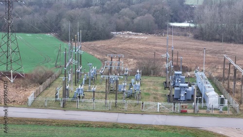 A fenced rural electrical substation surrounded by power lines, transformers, open agricultural fields and distant forest under an overcast sky, expressing industrial presence in natural countryside.