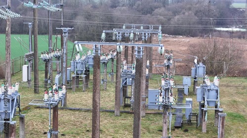 A fenced rural electrical substation surrounded by power lines, transformers, open agricultural fields and distant forest under an overcast sky, expressing industrial presence in natural countryside.