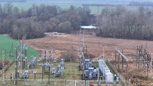 A fenced rural electrical substation surrounded by power lines, transformers, open agricultural fields and distant forest under an overcast sky, expressing industrial presence in natural countryside.