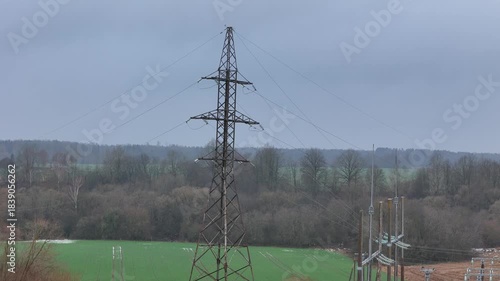 A fenced rural electrical substation surrounded by power lines, transformers, open agricultural fields and distant forest under an overcast sky, expressing industrial presence in natural countryside.