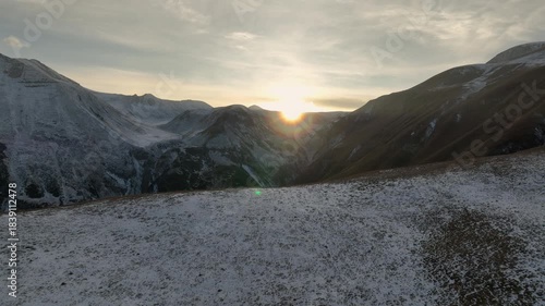 Aerial view of Majestic landscape of the Caucasus mountains at sunset. The sun shines brightly over snow-covered peaks, creating a dramatic lens flare and golden light over the valley.