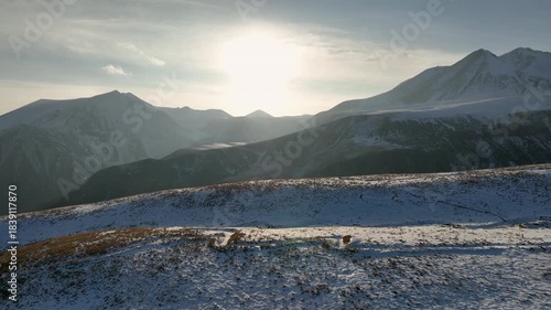 Aerial view of Majestic landscape of the Caucasus mountains at sunset. The sun shines brightly over snow-covered peaks, creating a dramatic lens flare and golden light over the valley.