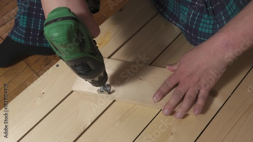Person using green cordless drill to fasten metal bracket to wooden plank lying on light wood surface, other hand holding board steady while kneeling in plaid pants.