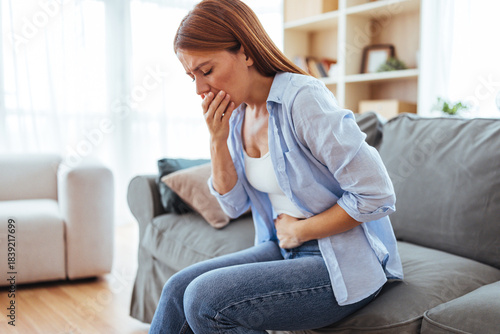 Woman Sits on Couch at Home Suffering Nausea and Stomach Discomfort, Gripping Her Abdomen