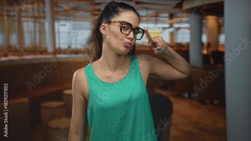 Woman wearing glasses and green tank top making peace sign with fingers in restaurant seating area; playfulness.