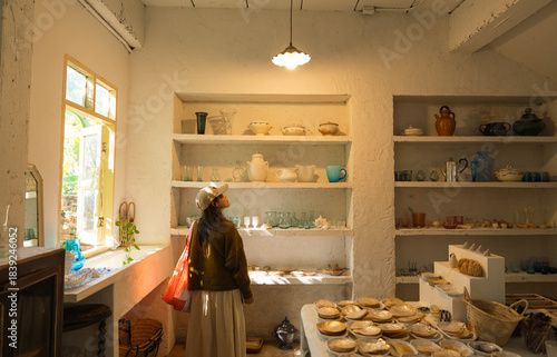 Woman in shop admires pottery and glass on shelves light streaming through the window