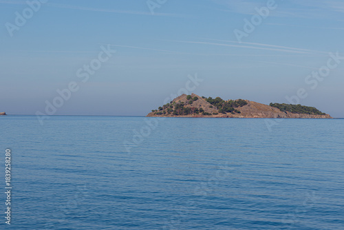 A serene island with sparse vegetation surrounded by calm blue sea under a clear sky.