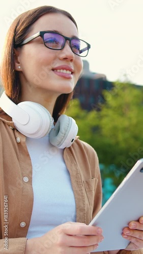 Portrait of young woman in city park with headphones around neck and glasses, working on tablet on commercial building background, outdoors