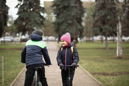 Cute little girl child and teenager boy rides an scooter and bike together in a park