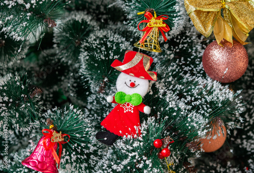 Festive Christmas ornaments hanging on a snowy pine tree. Glittery red and silver baubles, a gold and red bow, a snowman and a bell decorate the frosted green branches.