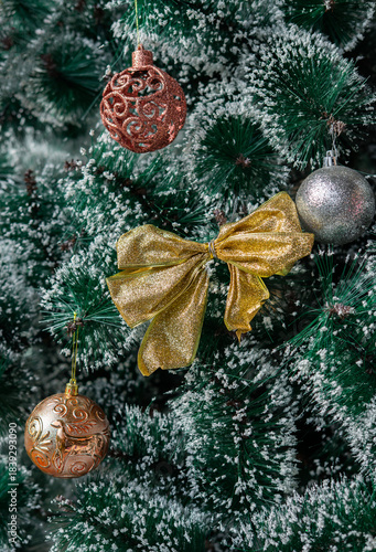 Festive Christmas ornaments hanging on a snowy pine tree. Glittery red and silver baubles, a gold and red bow, a snowman and a bell decorate the frosted green branches.