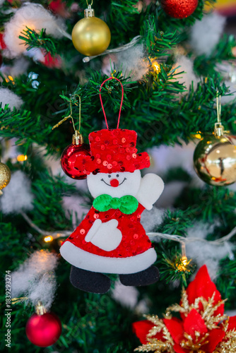 Festive Christmas ornaments hanging on a snowy pine tree. Glittery red and silver baubles, a gold and red bow, a snowman and a bell decorate the frosted green branches.