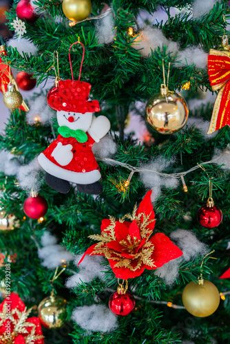 Festive Christmas ornaments hanging on a snowy pine tree. Glittery red and silver baubles, a gold and red bow, a snowman and a bell decorate the frosted green branches.
