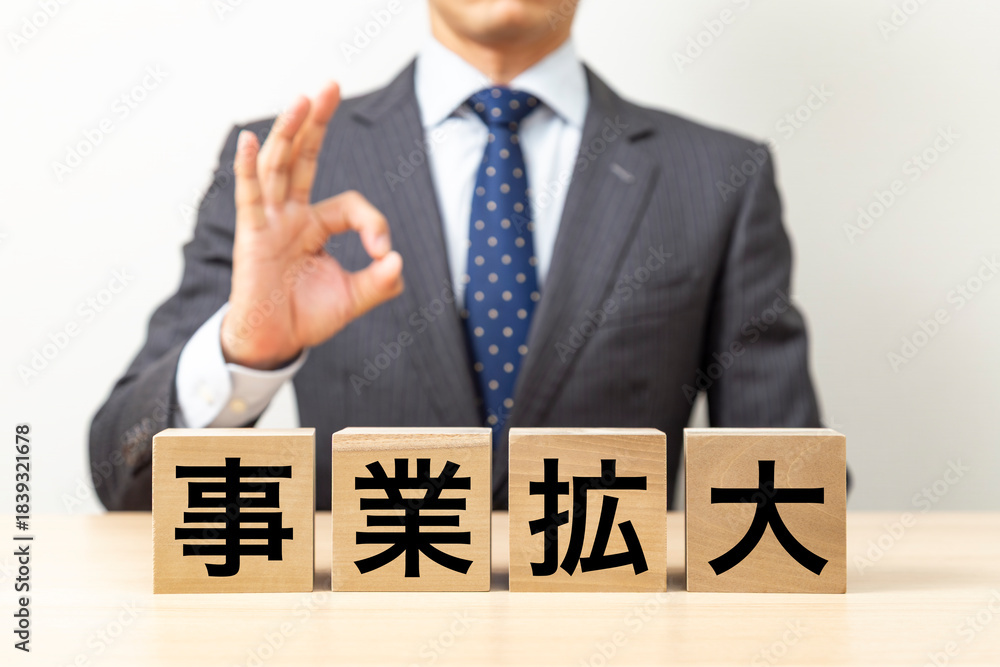 Obraz premium 「事業拡大」と書かれたブロックとOKポーズをするビジネスマン
