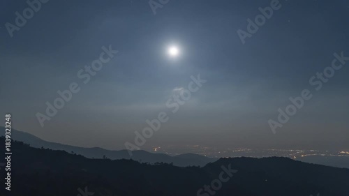 A bright full moon shines over a distant city in a dark valley, framed by rolling mountain ridges in Asia. Stunning night scene captured during a cold twilight for travel and celestial photography