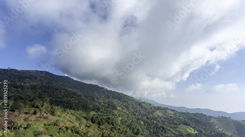 A vast billowing cloud passes over a steep, lush, green mountain slope with tropical forest in Asia. Stunning daytime natural scene captured during a clear day for environment and travel photography