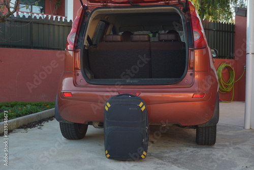 Suitcase standing on the ground behind an open car trunk, symbolizing the start of a journey or preparation for travel.