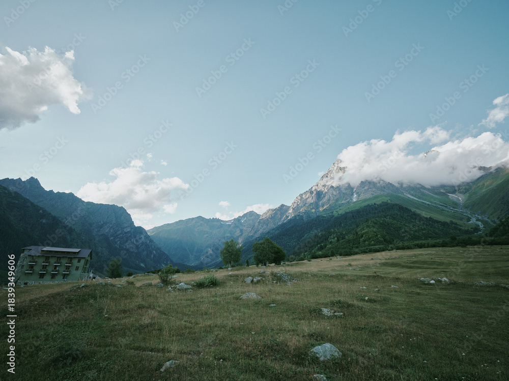 Fototapeta premium Mountain landscape, meadow, building, valley, clouds, sky, rural, grass, nature, scenery, expansive hillside view with distant peaks and a lone structure for exploration and photography adventure