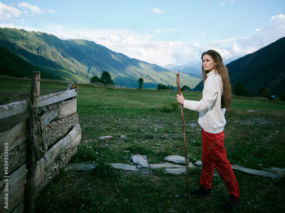 Fototapeta premium Hiking woman walks with a wooden staff across a alpine meadow toward distant mountains, rustic fence and wide sky, a quiet outdoor journey through open countryside