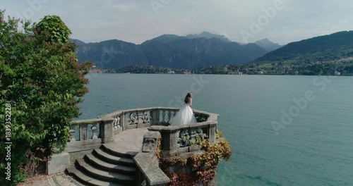 High-angle aerial drone shot of a bride in a white wedding dress run on a stone bastion or turret extending into deep blue lake water. The area is bordered by a beautiful green park