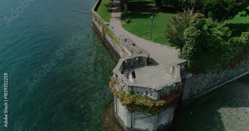 A high-angle drone view of a bride in a white wedding dress spinning on a stone turret or platform built into a retaining wall by deep blue lake water.