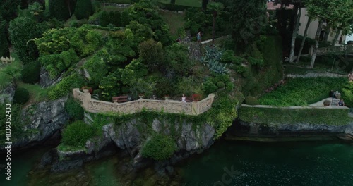 Expansive drone shot showcasing the historic Villa del Balbianello on its prominent peninsula on Lake Como, Italy. The view highlights the luxury villa's classic architecture, lush, manicured gardens,