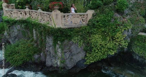 High-angle, near-vertical drone shot of a romantic wedding destination: a bride in a white gown stands on a small, ornate stone terrace with a carved railing, built into a cliff over rushing lake