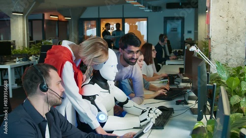 Humanoid robot typing on keyboard. Coworkers guiding task while smiling. People focusing on screens in active office. Team supporting robot during workflow. Shared workspace with group collaborating.