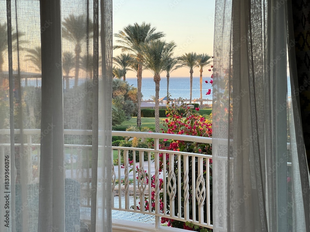 Obraz premium View from the room onto the balcony and the turquoise sea with the resort's palm trees at sunset. Travel destinations. Summer vacations. Year-round vacations