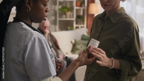 Medium shot of female medical worker in scrubs giving pills to middle-aged woman while prescribing medication to sick senior patient sitting in background at home
