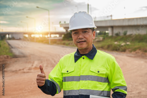 Construction Worker Wearing Safety Gear Giving Thumbs Up on Construction Site with Sunset in Background