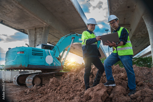 Engineers Collaborating on Construction Site Under Concrete Bridge with Heavy Machinery at Sunset