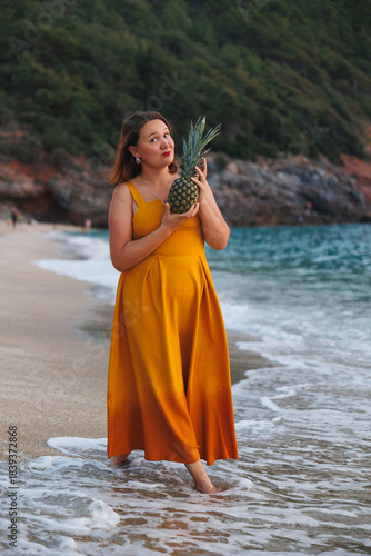 Woman By Shore Holding Pineapple Close, Wearing Vibrant Mustard Dress, Gentle Waves Lapping, Rocky Green