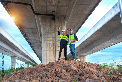 Two Construction Workers Inspecting Engineering Project Under Elevated Highway Structure at Dusk