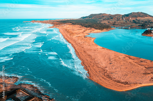 Drone shot of the Liencres coastline, revealing waves crashing along the sandbar while the serene blue estuary stretches inland under a clear sky.