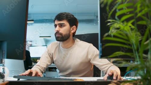 Close camera view of office worker rapidly typing. Male submitting work late while focusing on screen. People working in background at desks. Active office environment showing intense workflow.
