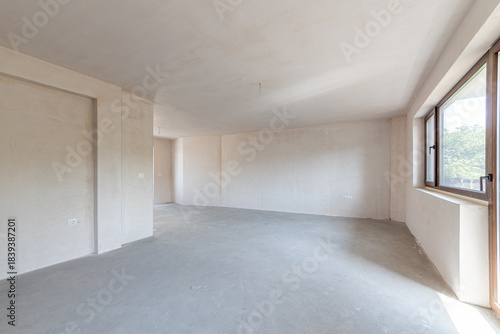 Interior of an empty room under construction in a new apartment. Unfinished space with raw concrete walls, floor, and a large window. Renovation and real estate concept.