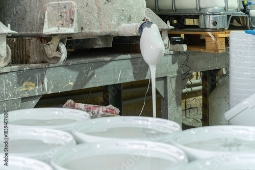 Manual paint pouring process in industrial workshop. White pigment flowing from industrial spout into containers during manual paint production in factory.