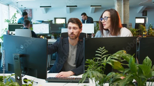 Caucasian female colleague standing beside desk pointing at monitor showing mistake in code. Male focusing on screen while receiving help. Modern office workspace with bright lights and many coworkers