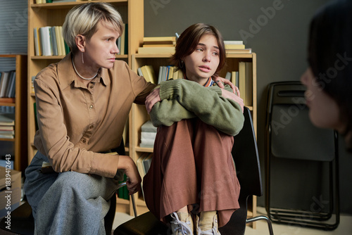 Adult woman comforting teenage girl sitting with knees to chest during group therapy session