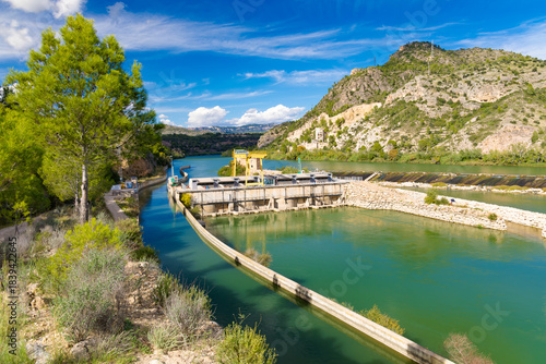 Scenic view of the Xerta Weir (Azud de Xerta) on the Ebro river in Catalonia, Spain, featuring a dam, green water, and rocky hills under a blue sky.