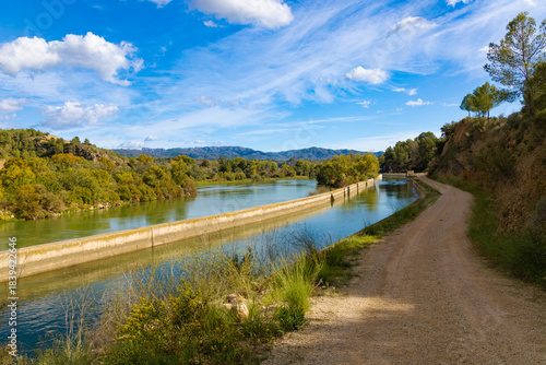 Water canal running alongside the Ebro river near Xerta Weir in Catalonia, Spain, with a dirt path and green trees under a blue sky.