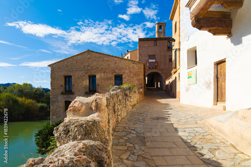 Cobblestone street ascending to Miravet castle with stone houses and Ebro river view in Catalonia, Spain, under a blue sky.