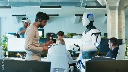 Male engineer giving voice instructions to humanoid robot. Man holding tablet while noting details. Robot raising hands as part of command test. In active tech office with focused staff.