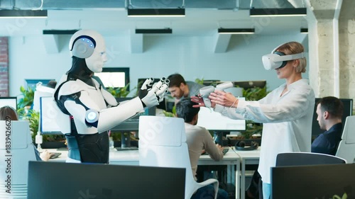 Female engineer guiding humanoid with VR controls. Woman raising hands while robot mirroring gesture. Motion sync test unfolding smoothly as staff continue working. Spacious tech office.