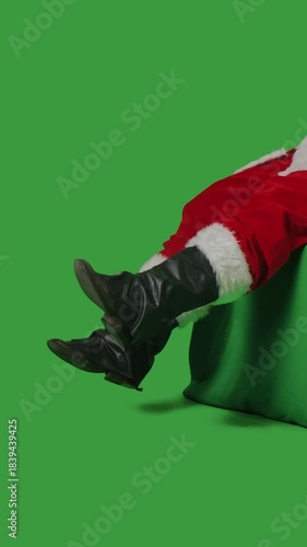 Santa Claus wiggles his feet while sitting on a green mockup against a green chroma key background