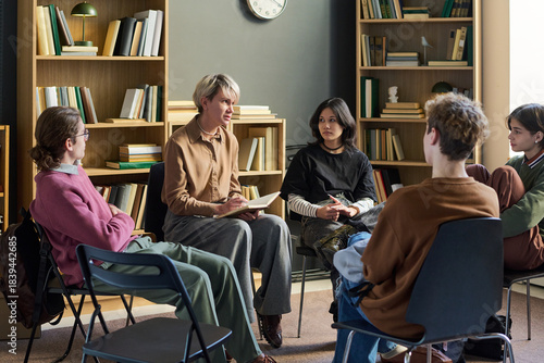 Group of diverse young people and teenagers sitting in circle participating in group therapy session, woman leading discussion while others listening attentively in library setting
