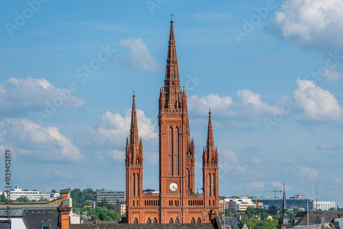 A telephoto shot of Wiesbaden Evangelical Market church on a clear spring day, bathed in sunlight against a backdrop of blue skies and white clouds.